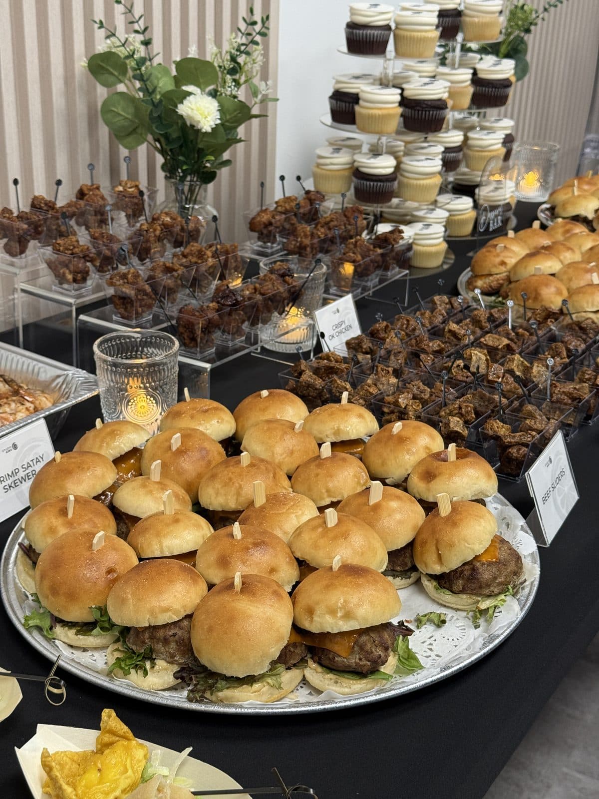 A table of sliders and shrimp satay skewers with tiers of cupcakes behind