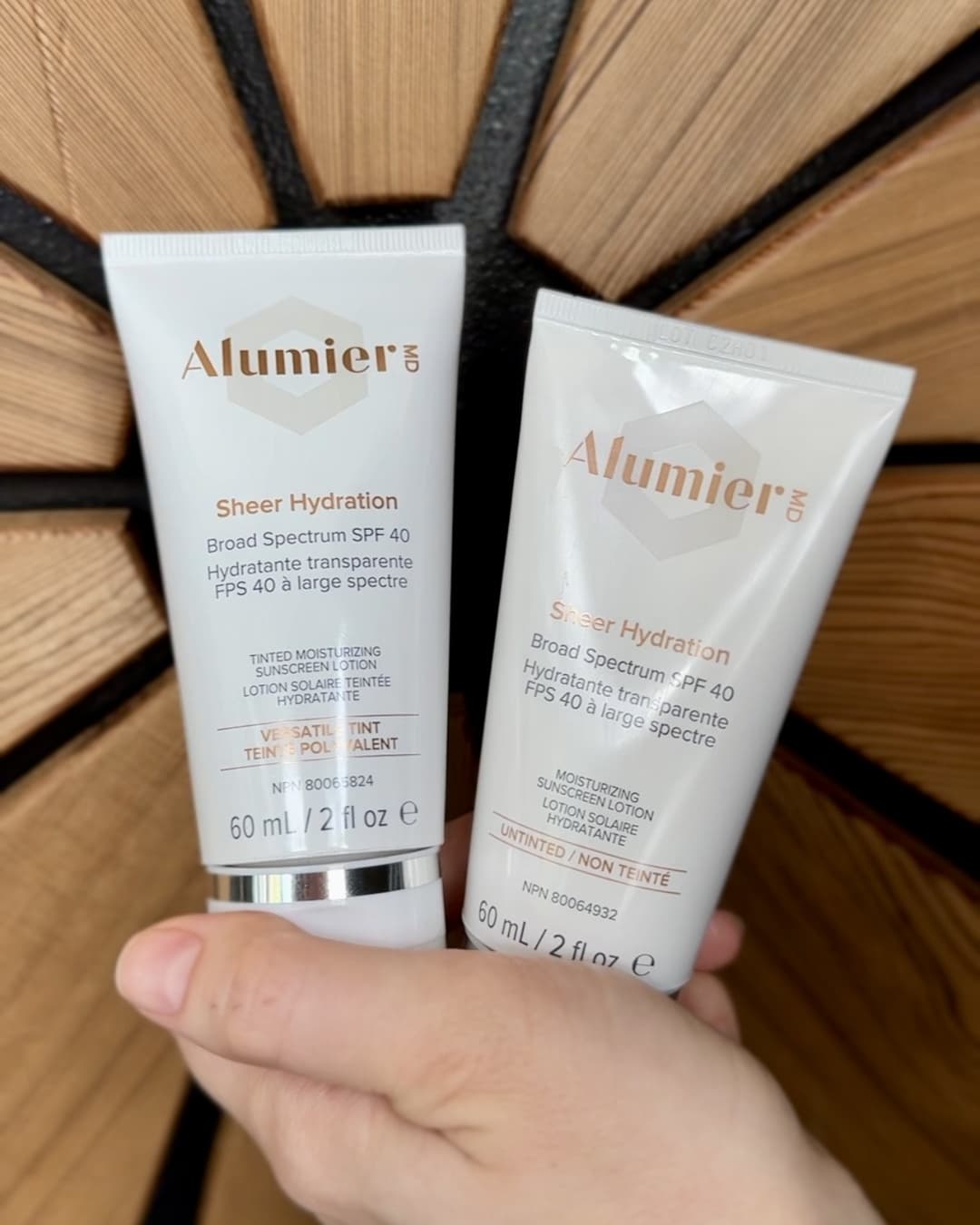 Two tubes of AlumierMD Sheer Hydration Broad Spectrum SPF 40 sunscreen, one tinted and one untinted, held in a hand against a wooden background