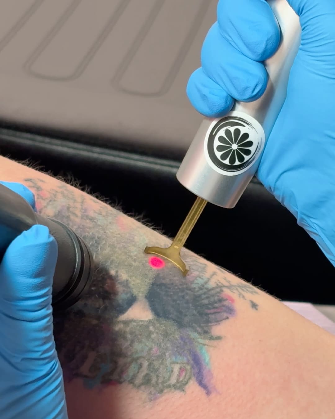 Studio Kiku technician using the PicoWay laser handpiece on a colourful arm tattoo, with the red aiming beam visible on the skin