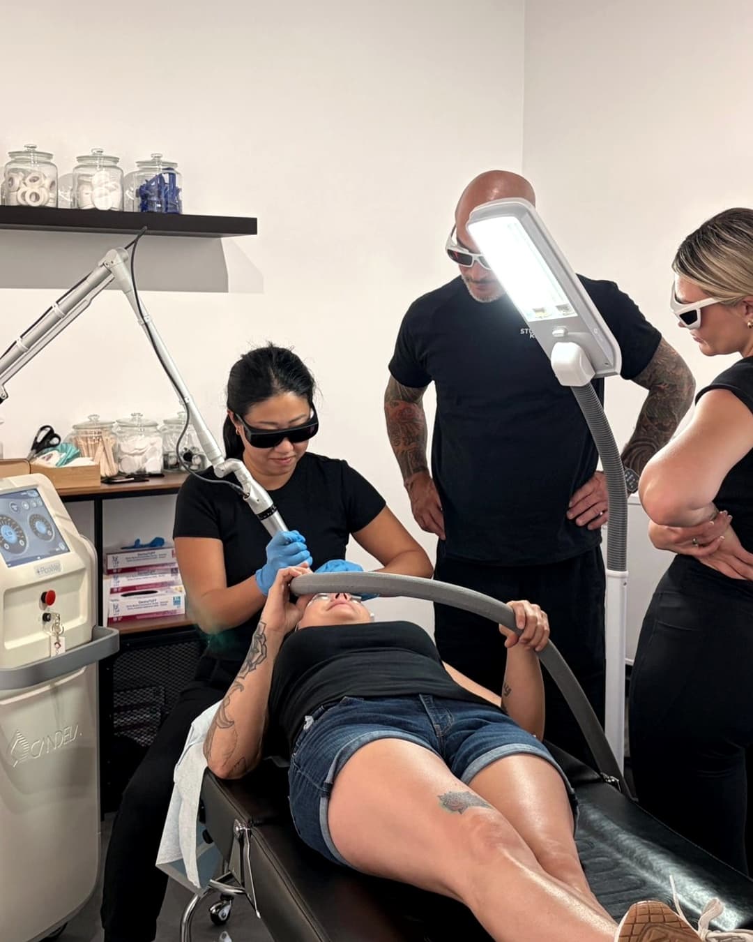 A Studio Kiku laser tattoo removal training session in progress, with a lead technician treating a live model on the PicoWay while Billy DeCola and a student observe
