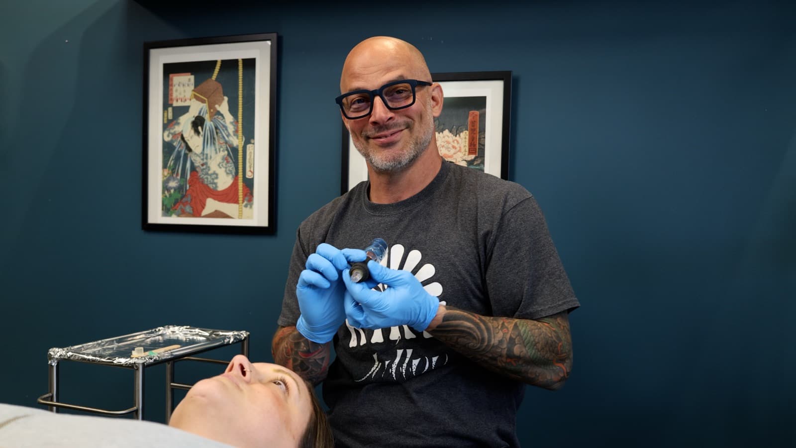 Billy DeCola holding a vial of saline solution beside a reclined client at Studio Kiku, before a permanent makeup removal session